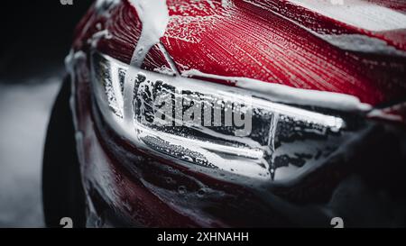 Nahaufnahme eines modernen Red Performance Car in einer dunklen Studio-Umgebung. Kreative LED-Scheinwerfer Foto eines Performance-Sportwagens, der während einer detaillierten Autowäsche in Shampoo tropft Stockfoto