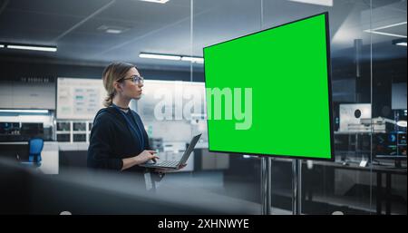 Wunderschöner leitender Software-Engineer, der allein in einem Konferenzraum steht, einen Laptop verwendet und einen Fernseher mit Green Screen Mock Up Display mit potenziellen Informationen ansieht, die von der Entwicklungsabteilung erstellt wurden Stockfoto