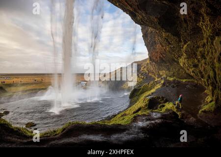 Island, Sudurland, eine Figur mit blauer Jacke und rosa Mütze in der Höhle des Seljalandsfoss Wasserfalls, 60 m hoch, denkt an einen von Stockfoto