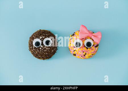 Zwei süße Kekse mit bunten Streuseln mit guten Augen und einer mit rosa Haarschleife auf hellem pastellblauem Hintergrund. Lustiges süßes Essen-Konzept. Stockfoto