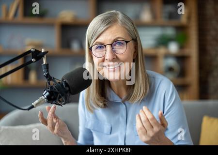 Lächelnde Seniorin nimmt Podcast aus dem Heimstudio auf. Begeisterter, reifer Podcaster, der Wissen und Erfahrungen teilt. Komfortable und professionelle Einrichtung. Ansprechende Kommunikation bei der Inhaltserstellung. Stockfoto