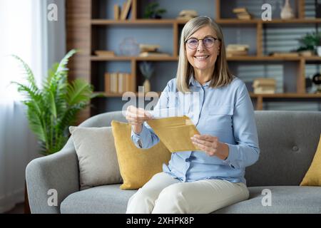 Glückliche ältere Frau, die auf grauem Sofa im gemütlichen Wohnzimmer sitzt und mit Freude einen Brief öffnet. Sie trägt ein blaues Hemd und eine Brille, umgeben von Bücherregalen und Zimmerpflanzen. Stockfoto