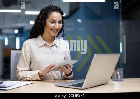 Lächelnde Geschäftsfrau mit Headset, die im modernen Büro auf einem Tablet arbeitet. Professionelle Frau, die mit einem Laptop interagiert und Technologie- und Kommunikationsfähigkeiten demonstriert. Stockfoto