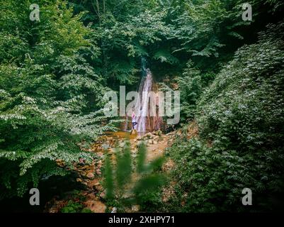 [So hoch wie der Himmel] Ein Mädchen allein zwischen den grünen Bäumen und vor dem Wasserfall erhält positive Energie von der Natur, indem es Yoga macht. Stockfoto