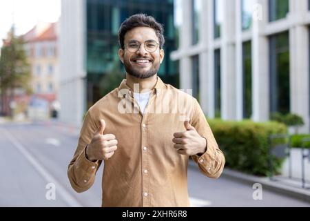 Ein lächelnder hispanischer Mann in lässigem Outfit, der die Daumen hoch auf der Straße der Stadt hält. Junge Profis, die positive Einstellung, Selbstvertrauen und Anerkennung in der städtischen Umgebung zeigen. Moderne Gebäude im Hintergrund. Stockfoto