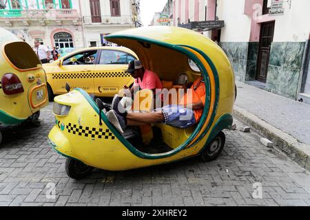 Havanna, Kuba, Zentralamerika, Ein gelbes, dreirädriges Taxi steht auf einer Kopfsteinpflasterstraße, ein Mann sitzt darin, umgeben von alten Gebäuden Stockfoto