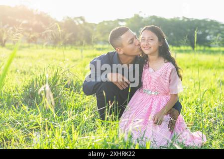 lateinischer Vater, der seiner kleinen brünetten Tochter die Wange küsst, während er auf dem Gras sitzt und einen Sonnenuntergang im Hintergrund hat Stockfoto