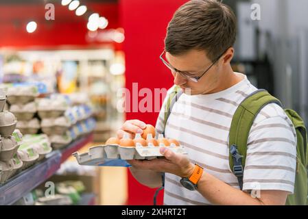 Der Mann inspiziert sorgfältig einen Karton mit Eiern im Gang des Lebensmittelgeschäfts und stellt vor dem Kauf sicher, dass diese Qualität gewährleistet ist. Mann kauft Lebensmittel im Supermarkt zum Frühstück. Stockfoto