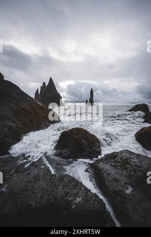 Felsen, die von Wellen am schwarzen Sandstrand von Reynisfjara auf Island an einem bewölkten und windigen Tag getroffen werden. Stockfoto