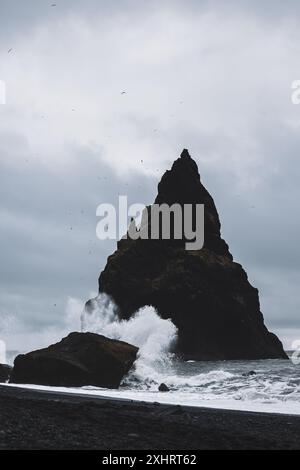 Blick auf eine riesige Felsformation am schwarzen Sandstrand von Reynisfjara in Island, der an einem bewölkten und windigen Tag von riesigen Wellen getroffen wird. Stockfoto