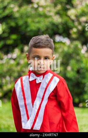 Porträt eines kleinen lateinjungen, der in rotem Abschlusskleid ohne Mütze in einem grünen Park posiert Stockfoto