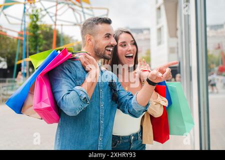 Ein junger erwachsener Mann zeigt auf die Ladenpräsentation eines Ladengeschäfts im Einkaufszentrum, während seine Freundin lächelt und denkt, sie würde etwas kaufen. Ein echtes Paar schlendert durch das Einkaufszentrum und hält Einkaufstaschen. Hochwertige 3D-Illustration Stockfoto