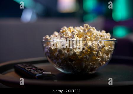Konzentrieren Sie sich auf eine Schale Popcorn auf dem Tisch neben der Fernbedienung mit neonbeleuchtetem Home Entertainment-Apartment in unscharfem Hintergrund. Nahaufnahme eines Snacks, der für den kommenden Filmabend im Wohnzimmer vorbereitet wurde Stockfoto