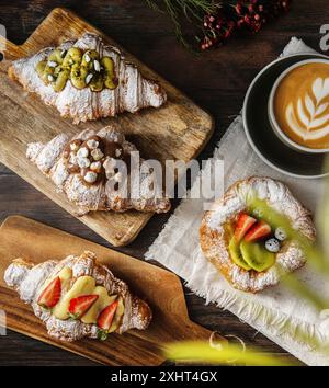 Frisch gebackene Gourmet-Croissants und Blätterteig mit Beeren, Marmelade, Schokolade und Pistazien. Cappuccino-Tasse auf dunklem Holzhintergrund. Draufsicht. Stockfoto