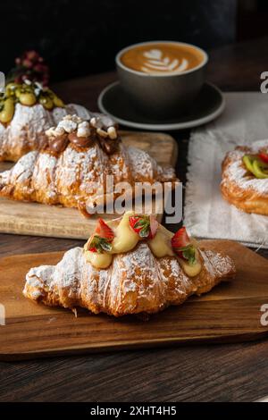 Frisch gebackene Gourmet-Croissants und Blätterteig mit Beeren, Marmelade, Schokolade und Pistazien. Cappuccino-Tasse auf dunklem Holzhintergrund. Stockfoto