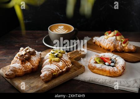 Frisch gebackene Gourmet-Croissants und Blätterteig mit Beeren, Marmelade, Schokolade und Pistazien. Cappuccino-Tasse auf dunklem Holzhintergrund. Stockfoto