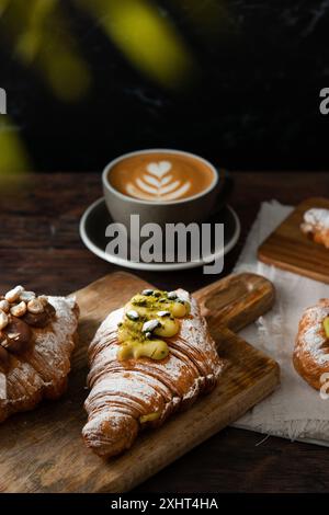 Frisch gebackene Gourmet-Croissants und Blätterteig mit Beeren, Marmelade, Schokolade und Pistazien. Cappuccino-Tasse auf dunklem Holzhintergrund. Stockfoto