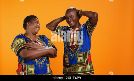 Fröhlicher Ehemann und Frau lachen zusammen vor der Kamera, haben Spaß mit albernen Witzen über orangem Hintergrund. afroamerikanisches Paar, das lustige Momente genießt, sorglose Leute lachen. Stockfoto