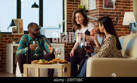 Freunde, die sich lebhaft unterhalten, Wein trinken und Snacks von der Wurstplatte zu Hause essen, handgeführte Kameraaufnahme. BIPOC-Kollegen entspannen sich im modernen Wohnraum des Apartments und genießen das Wochenende Stockfoto