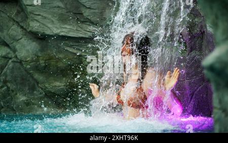 Eine Frau im Bikini steht unter einem Wasserfall, umgeben von spritzendem Wasser. Ihr Ausdruck ist heiter und fröhlich und fängt das Wesen der Freiheit ein Stockfoto