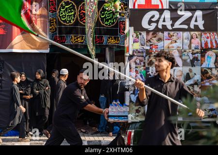 Kaschmiri schiitische Frauen beobachten, wie Trauernde mit palästinensischen Fahnen und Spruchbanner während einer Prozession am achten Tag von Muharram vorbeigehen. Im zweiten Jahr in Folge erlaubte die Regierung von Jammu und Kaschmir die 8. Muharram-Prozession vom Guru-Basar nach Dalgate in Srinagar. Trotz eines seit langem bestehenden Verbots hielten die schiitischen Muslime weiterhin Prozessionen auf dem 8. Und 10. Muharram entlang dieser Routen ab, die oft mit Polizeieingriffen konfrontiert waren. Die diesjährige Prozession war ein bedeutendes und friedliches Gedenken an das Martyrium von Hazrat Imam Hussainís (AS), das Tausende von Trauernden anzog. Die Prozedur Stockfoto