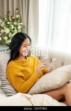 Eine glückliche, schöne asiatische Frau in kuscheliger Kleidung benutzt ihr Smartphone auf einer Couch im Wohnzimmer, chattet mit ihren Freunden oder scrollt in sozialen Netzwerken Stockfoto