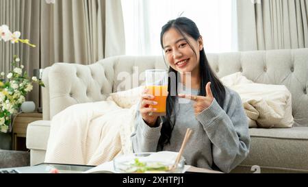 Eine attraktive, lächelnde junge asiatische Frau zeigt mit dem Finger auf ein Glas Orangensaft in der Hand, während sie an einem Couchtisch in der lebenden Wohnung sitzt Stockfoto