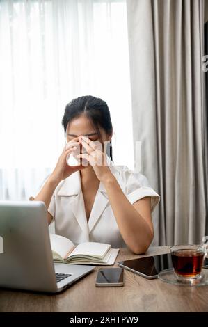 Eine gestresste, frustrierte asiatische Frau leidet unter Kopfschmerzen, denkt über Probleme nach, macht sich Sorgen um ihre Arbeit, berührt ihr Gesicht, sitzt an ihrem Schreibtisch Stockfoto