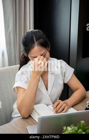 Eine gestresste, frustrierte asiatische Frau leidet unter Kopfschmerzen, denkt über Probleme nach, macht sich Sorgen um ihre Arbeit, berührt ihr Gesicht, sitzt an ihrem Schreibtisch Stockfoto