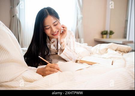 Eine schöne asiatische Frau im Pyjama schreibt ihr Tagebuch auf dem Bett im Schlafzimmer. Freizeit, Hobby, häusliches Leben Stockfoto