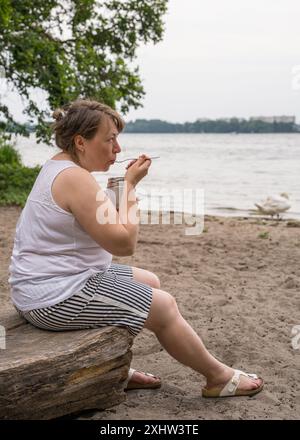 Eine übergewichtige Frau mittleren Alters ist draußen in der Nähe eines Sees und isst Nahrung aus einer Thermoskanne Stockfoto