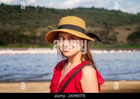 Mädchen mit Hut, das vor der Kulisse der Vögel Pelikane und Flamingos im See läuft. Glückliche junge Frau in rotem T-Shirt, die während der sonnigen Savanne erkundet Stockfoto