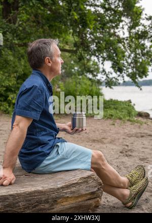 Ein Mann mittleren Alters reist, sitzt am Ufer des Sees, isst Nahrung aus einer Thermoskanne und blickt in die Ferne. Meditation, Genuss, Entspannung. Stockfoto