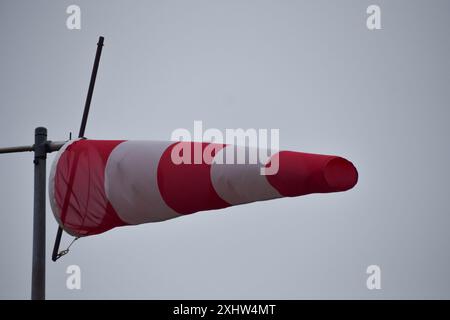Dieses Bild zeigt eine rot-weiße Windsocke, die vor einem grauen, bewölkten Himmel sichtbar ist. Die Windsocke, die auf einem Metallständer montiert ist, bietet einen cl Stockfoto