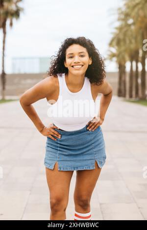 Porträt einer jungen lächelnden Frau mit den Händen auf den Hüften, die ihre Zeit in einem Stadtpark mit Palmen im Hintergrund genießt. Stockfoto