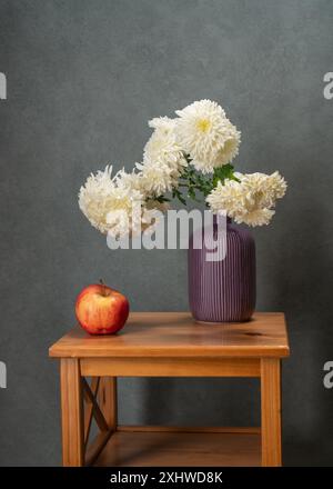 Viele weiße Blumen. Strauß weißer Astern in einer lila Vase auf einem Holztisch. Daneben steht ein roter Apfel. Grauer Hintergrund. Vitamine. Gesundheit. Stockfoto