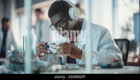 Porträt eines schwarzen männlichen Laborspezialisten beim Löten einer Leiterplatte mit Kopfhörern in einem Industrieunternehmen. Professioneller Mann, der Musik hört, arbeitet an einem technologischen Projekt für ein Startup Stockfoto