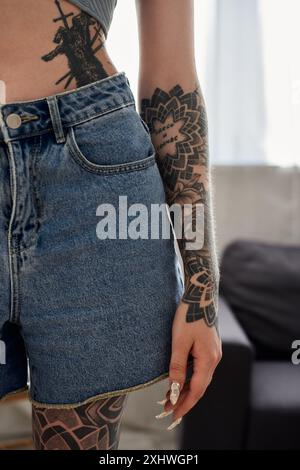 Eine tätowierte Frau entspannt sich zu Hause in ihrer modernen Wohnung, trägt Jeansshorts und ein lässiges Oberteil. Stockfoto