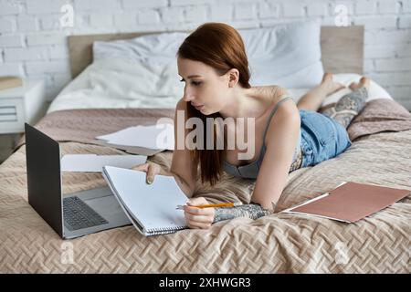 Eine junge Frau mit Tattoos arbeitet von zu Hause aus auf ihrem Laptop, während sie auf einem Bett liegt. Stockfoto