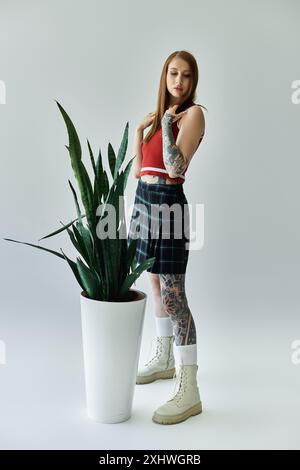 Eine tätowierte junge Frau, die einen karierten Rock und ein rotes Tanktop trägt, steht neben einer großen grünen Pflanze in einem weißen Topf. Stockfoto