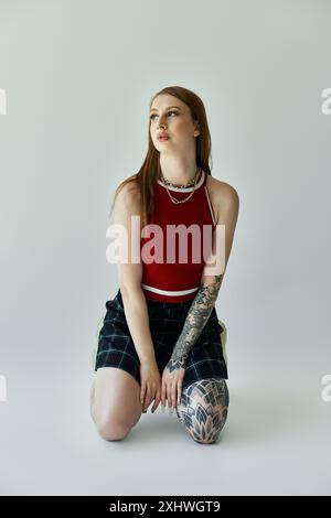 Eine junge Frau mit Tätowierungen posiert in einem roten Tanktop und einem karierten Rock vor grauem Hintergrund. Stockfoto