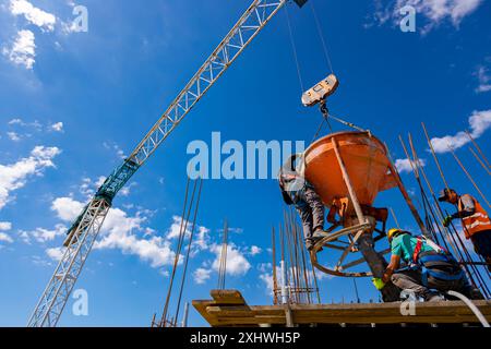 Bauarbeiter entladen konische Metallschaufeln, transportieren und liefern Zementgemische hoch und gießen Beton in eine hohe vertikale Form für den Bau Stockfoto