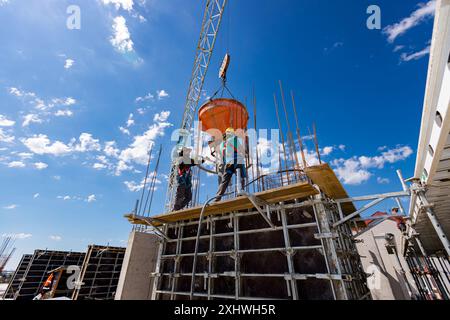 Bauarbeiter entladen konische Metallschaufeln, transportieren und liefern Zementgemische hoch und gießen Beton in eine hohe vertikale Form für den Bau Stockfoto