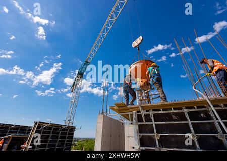 Bauarbeiter entladen konische Metalleimer, gießen Beton in eine hohe vertikale Form für die Säulen des Gebäudes und verwenden einen Kran, während er den Kegel t trägt Stockfoto