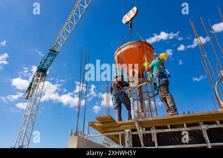 Bauarbeiter entladen konische Metallschaufeln, transportieren und liefern Zementgemische hoch und gießen Beton in eine hohe vertikale Form für den Bau Stockfoto