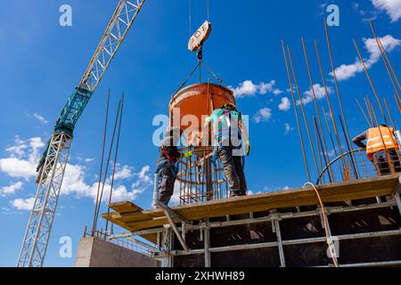 Bauarbeiter entladen konische Metalleimer, gießen Beton in eine hohe vertikale Form für die Säulen des Gebäudes und verwenden einen Kran, während er den Kegel t trägt Stockfoto