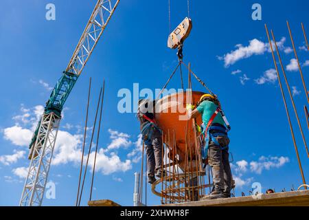 Bauarbeiter entladen konische Metallschaufeln, transportieren und liefern Zementgemische hoch und gießen Beton in eine hohe vertikale Form für den Bau Stockfoto