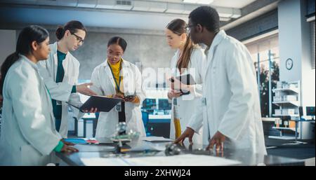 Ingenieure in Laborbeschichtungen mit Laptop und Tablet, Ideenfindung für elektronische Komponenten. Verschiedene Teams von Spezialisten für industrielle Robotik versammelten sich an einem Tisch, um einen Blueprint zu besprechen. Stockfoto