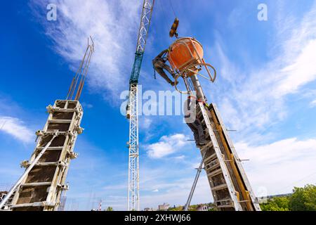 Bauarbeiter entladen konische Metallschaufeln, transportieren und liefern Zementgemische hoch und gießen Beton in eine hohe vertikale Form für den Bau Stockfoto