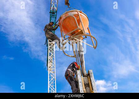 Bauarbeiter entladen konische Metallschaufeln, transportieren und liefern Zementgemische hoch und gießen Beton in eine hohe vertikale Form für den Bau Stockfoto